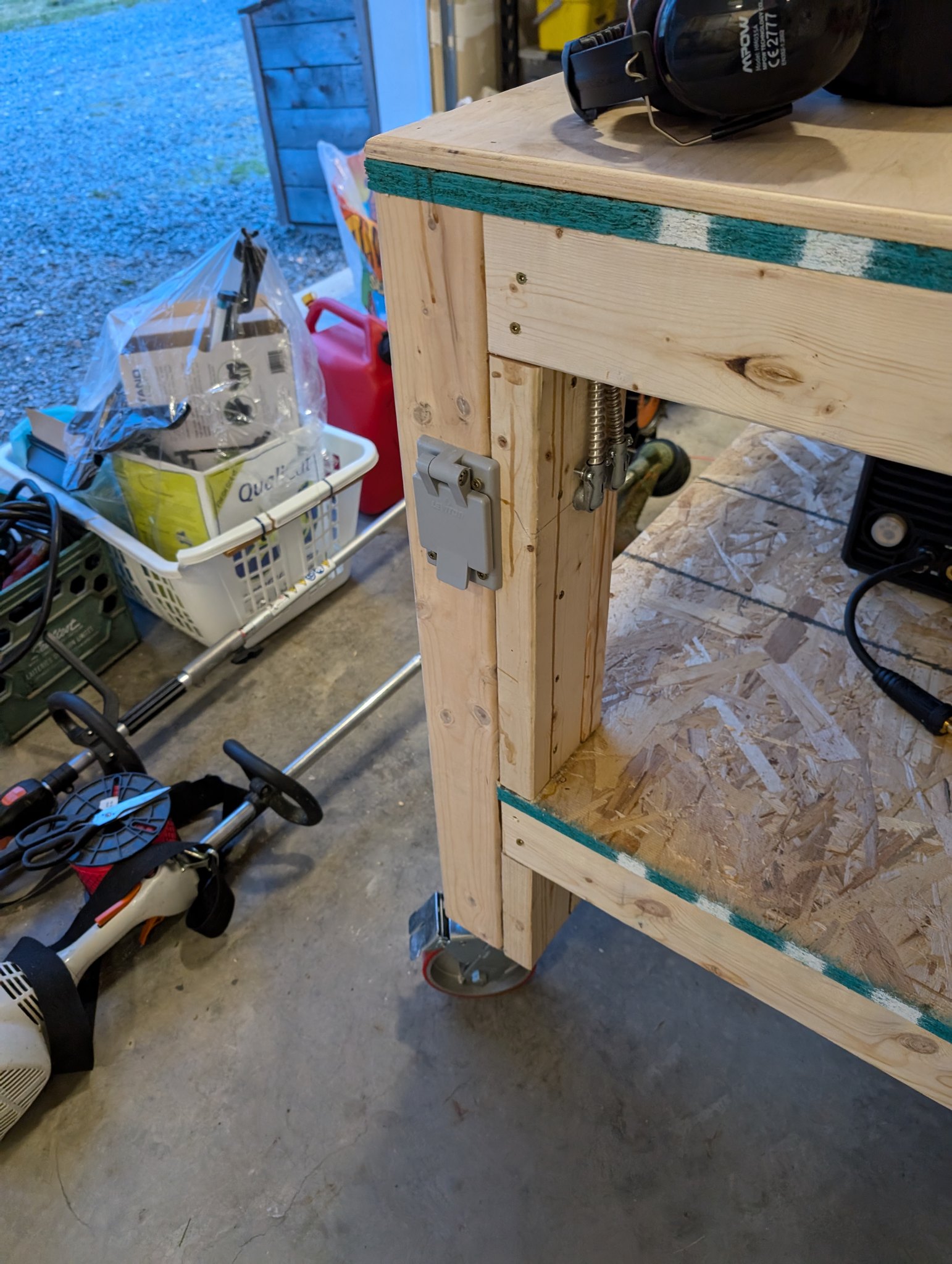 Covered electrical outlet mounted on a workbench leg beside metal conduit and the lower shelf